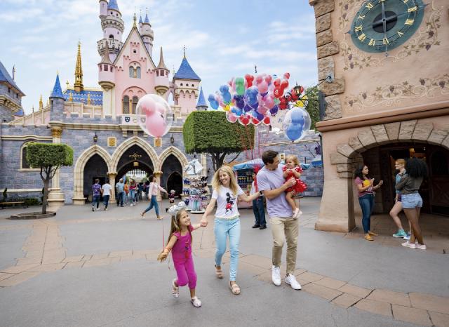 A family walks hand in hand near Sleeping Beauty Castle at Disneyland Park. The mother holds a child’s hand while the father carries another child. Colorful Mickey balloons float in the background, capturing the magic of Fantasyland.