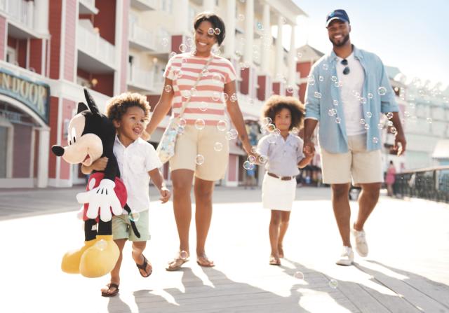 A happy family walking along Disney’s BoardWalk Inn, with a young child holding a Mickey Mouse plush and bubbles floating around.