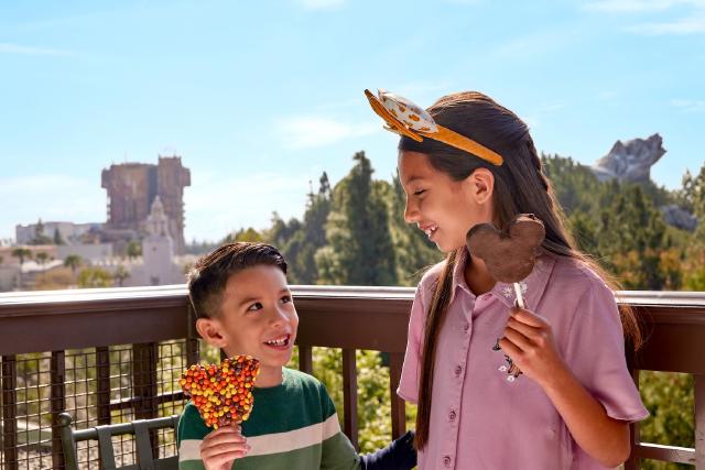 Two children smile at each other while enjoying Mickey-shaped treats at Disney's Grand Californian Hotel & Spa. The Guardians of the Galaxy – Mission: BREAKOUT! attraction and Grizzly Peak at Disney California Adventure Park are visible in the background.