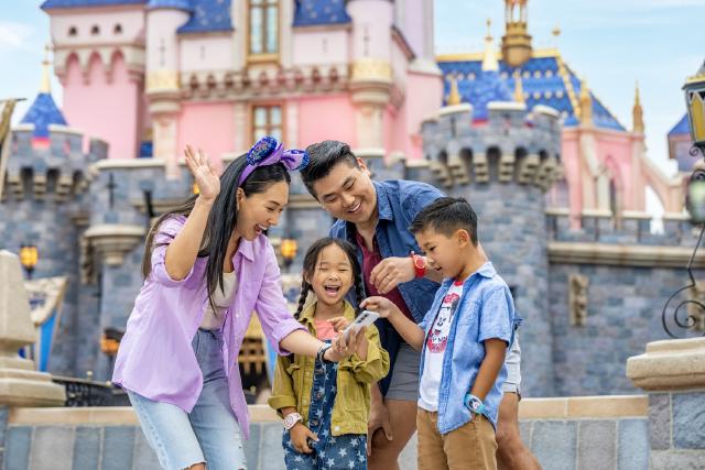 A family smiles and gathers around a smartphone using the Disneyland app in front of Sleeping Beauty Castle at Disneyland Park. The parents and children wear Disney-themed outfits and MagicBands.