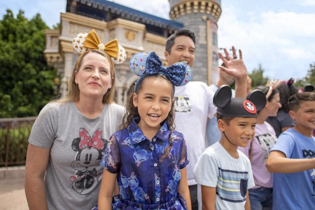 A family and children smile while enjoying a parade at Magic Kingdom Park at Walt Disney World Resort. They wear Disney-themed outfits and Mickey ears, with Cinderella Castle in the background.