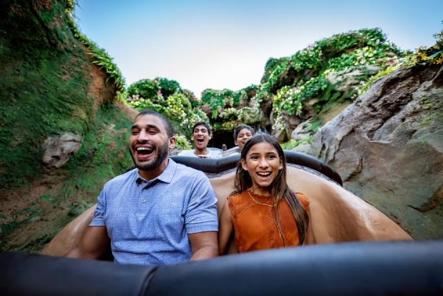 A family smiles and laughs while riding Tiana's Bayou Adventure, now at Magic Kingdom Park and Disneyland Park. Lush greenery and flowers surround the log flume as they enjoy the thrilling journey.