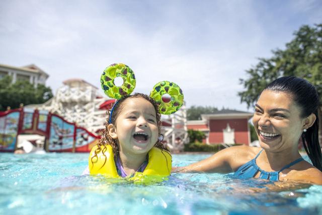 A mother and child smile while enjoying the Luna Park Pool at Disney’s BoardWalk Inn. The 1920s amusement park-inspired pool features a carnival theme, with a 200-foot-long waterslide and vibrant red and white decor in the background.