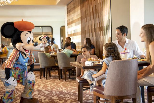 A family enjoys breakfast at Topolino’s Terrace – Flavors of the Riviera at Disney’s Riviera Resort. Mickey Mouse, dressed as an artist, greets guests, while Minnie Mouse appears in the background.