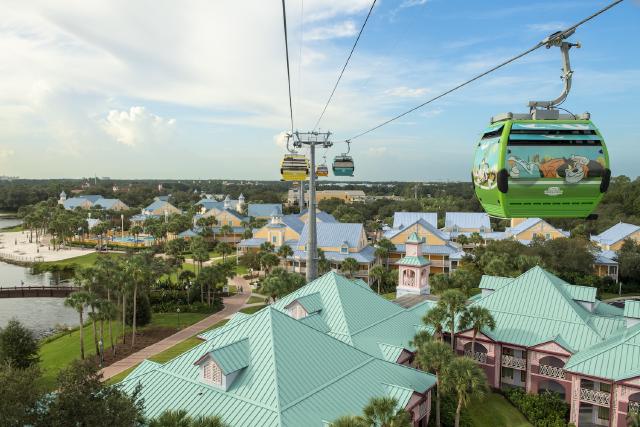 The Disney Skyliner gondolas glide above Disney’s Caribbean Beach Resort, offering a scenic view of the vibrant, tropical-themed buildings and lush landscapes below.