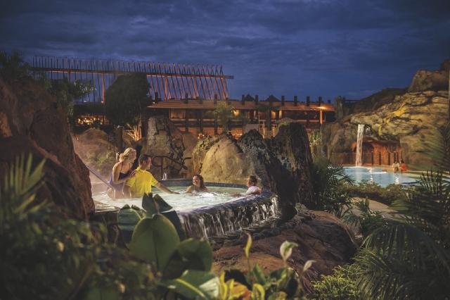 Guests relax in a themed hot tub at Disney’s Polynesian Village Resort. Lush greenery, rock formations, and a waterfall create a serene nighttime atmosphere.