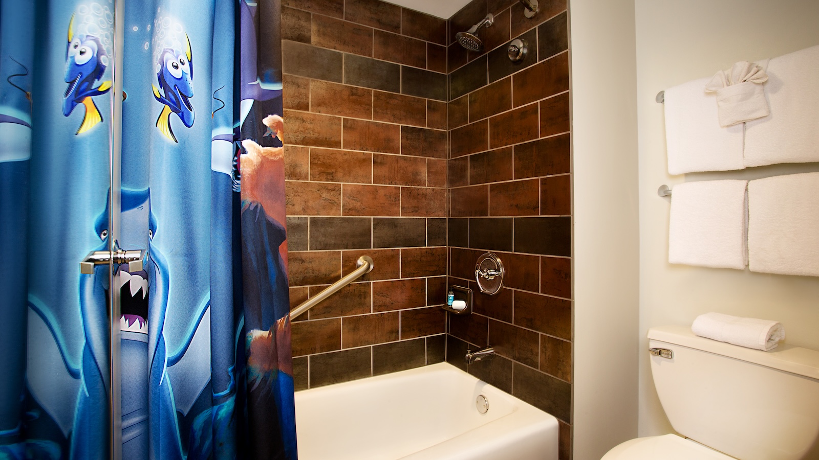 Bathroom at Disney’s Art of Animation Resort Finding Nemo Family Suite featuring a tub with brown tiled walls, a Finding Nemo-themed shower curtain, and neatly folded white towels on wall racks.