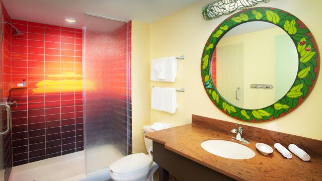 Bathroom at Disney’s Art of Animation Resort The Lion King Family Suite featuring a walk-in shower with red sunset-themed tiles, a green leaf-patterned mirror, and neatly arranged white towels on wall racks.