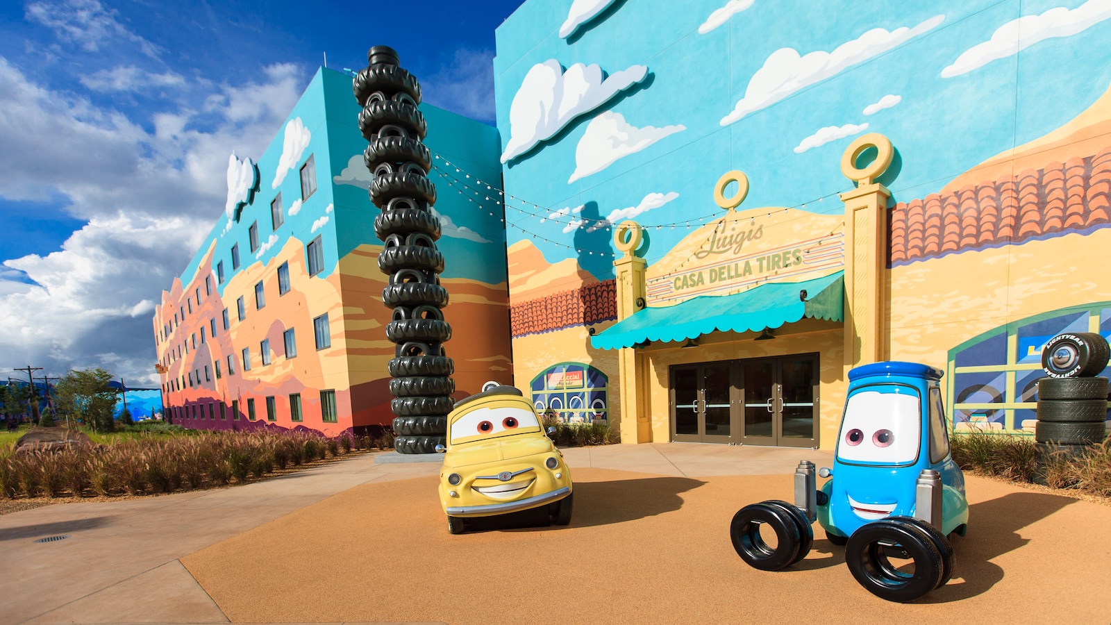 Cars-themed courtyard at Disney’s Art of Animation Resort featuring colorful building murals, a tower of tires, Luigi’s Casa Della Tires facade, and life-sized sculptures of Luigi and Guido under a bright blue sky.