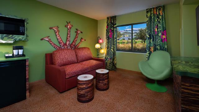 Living area of Disney’s Art of Animation Resort The Lion King Family Suite featuring a sofa with giraffe-themed decor, two wooden-style stools, a kitchenette with a microwave and mini-fridge, and a window showing outdoor The Lion King-themed scenery.