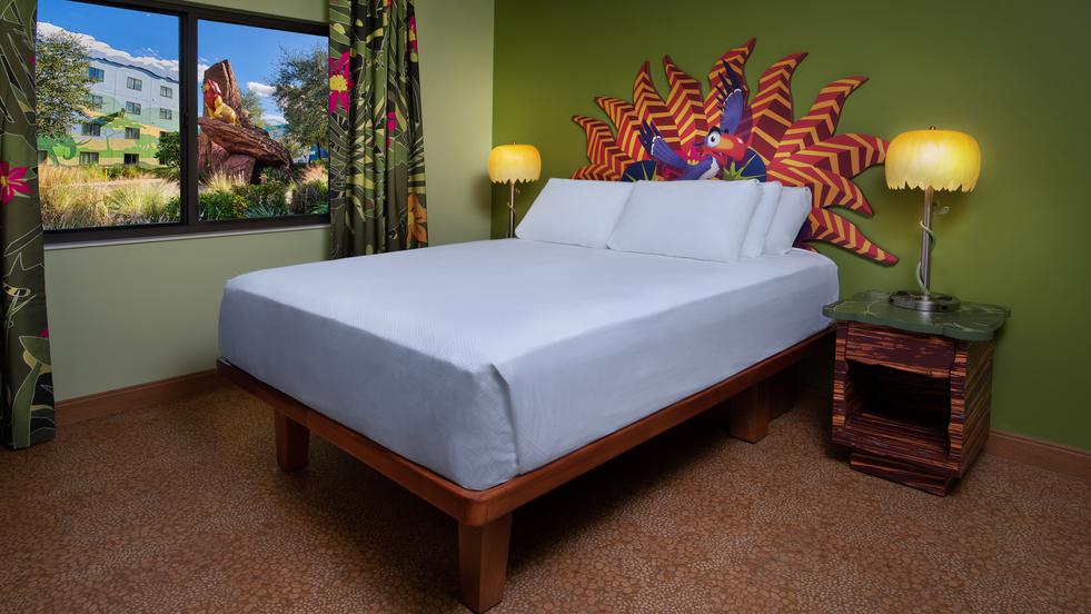 Brightly themed The Lion King Family Suite at Disney’s Art of Animation Resort featuring a queen bed with a colorful headboard, green walls, and yellow lamp accents. A window shows Mufasa on Pride Rock from The Lion King outside.